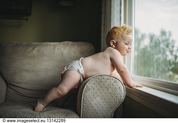 Shirtless cute baby boy looking through window at home