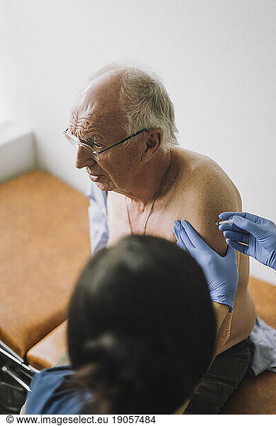 Shirtless male senior patient taking vaccination on arm at hospital