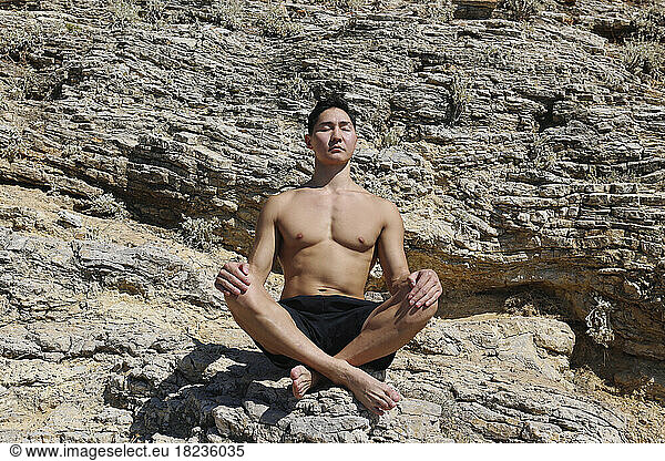 Shirtless young man with eyes closed meditating on rock at sunny day