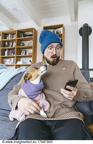 Shocked man with wooly hat sitting on couch holding dog checking smartphone