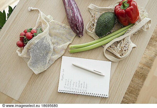 Shopping list by fresh vegetables in reusable bags on table at home