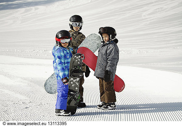 Siblings carrying snowboards while standing on snow covered field