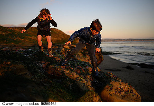 Siblings climbing down rocks near the ocean at low tide and sunset