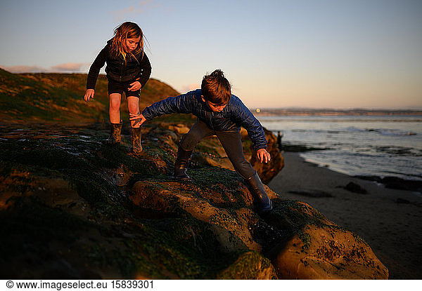 Siblings climbing on the rocks near the ocean at low tide and sunset