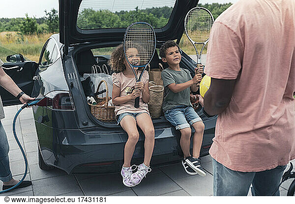 Siblings holding tennis rackets while sitting in car trunk and looking at father