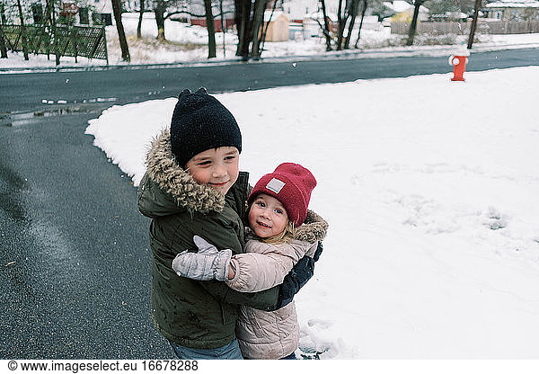 Siblings hugging each other in their driveway while playing in snow.