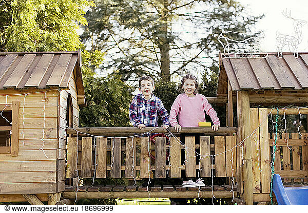 Siblings standing on wooden footbridge by playhouse in back yard