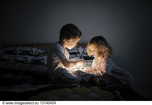 Siblings with illuminated string lights sitting on bed at home