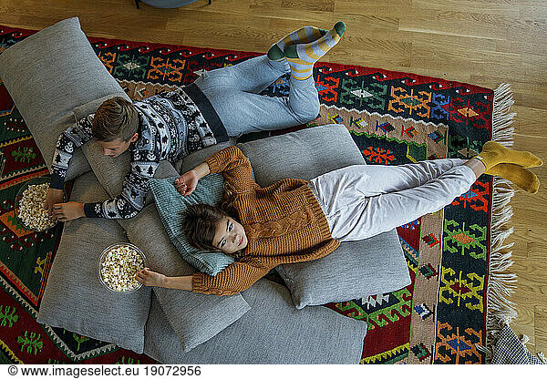 Siblings with popcorn bowls lying on pillows at home