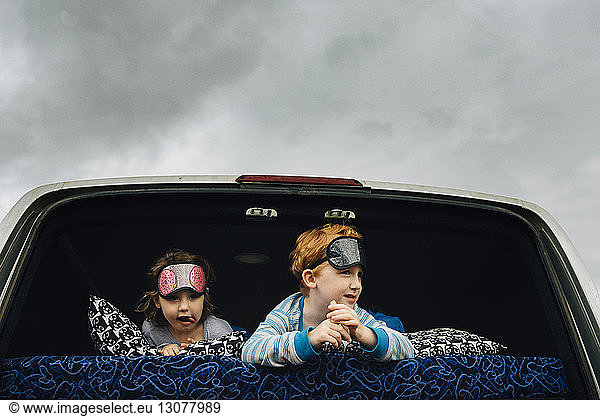Siblings with sleep masks lying in vehicle while looking away against cloudy sky