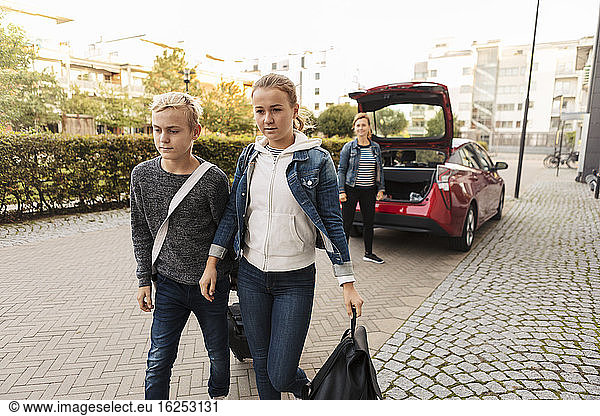 Siblings with suitcase walking while smiling mother standing by car on road
