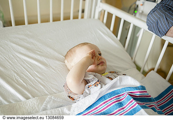 Sick baby boy rubbing eye while lying in hospital crib