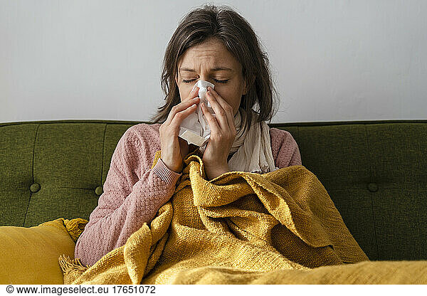 Sick woman blowing nose in facial tissue sitting on sofa at home