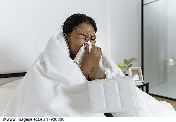 Sick woman sitting on bed wrapped in a blanket blowing nose in a tissue