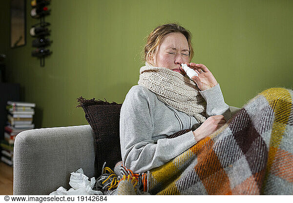Sick young woman using nasal spray relaxing on couch
