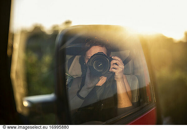 Side-view mirror reflection of woman photographing setting sun