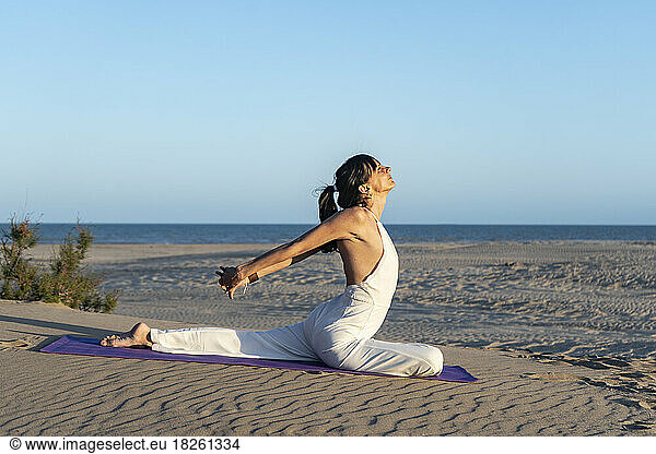 Side view of a woman practising yoga on a mat on the beach