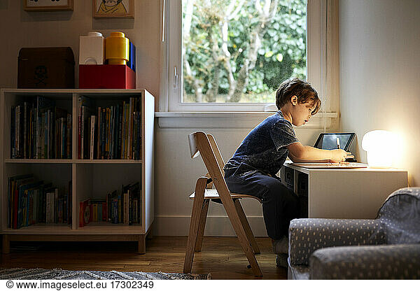 Side view of boy drawing while sitting on chair by window at home