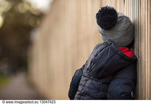 Side view of boy wearing warm clothing leaning on wooden fence in backyard