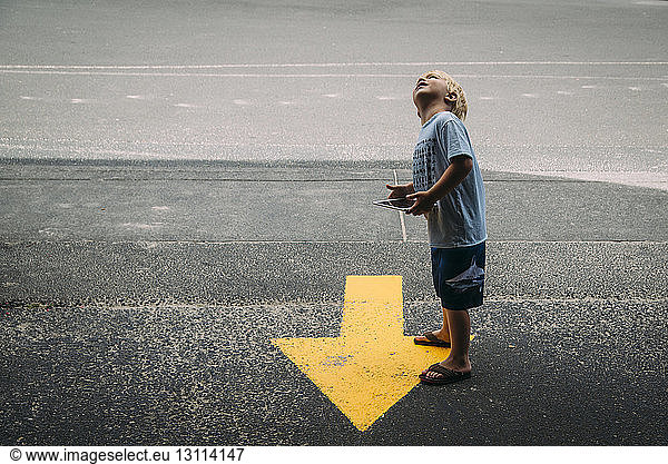 Side view of boy with tablet computer looking up while standing by arrow symbol on road