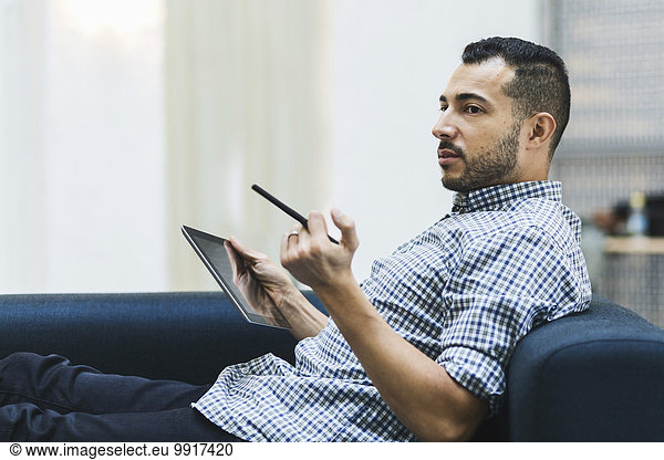 Side view of businessman holding digital tablet while relaxing on sofa at office