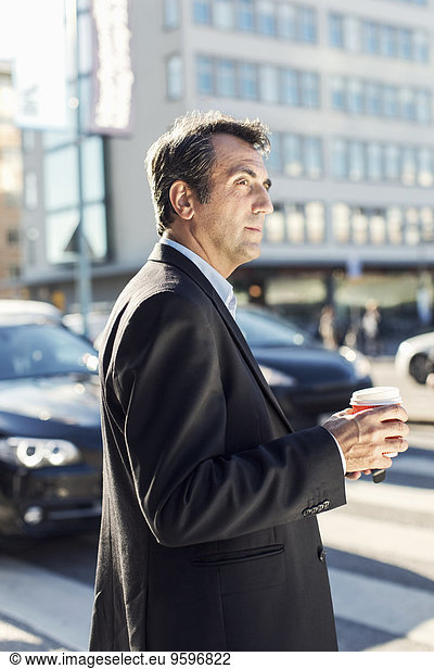 Side view of businessman looking away while holding disposable coffee cup in city