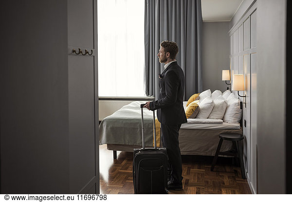 Side view of businessman standing with luggage by bed in hotel room