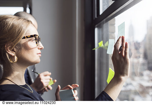 Side view of businesswoman sticking notes on glass window at office