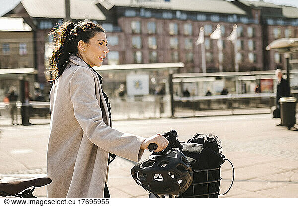 Side view of businesswoman wearing long coat while holding bicycle in city