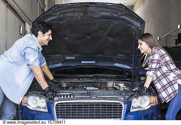 Side view of cheerful casual man and woman standing at car with opened bonnet and looking at each other working in team in Madrid