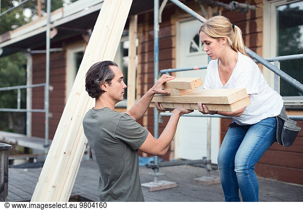 Side view of couple carrying stack of lumber outside house being renovated