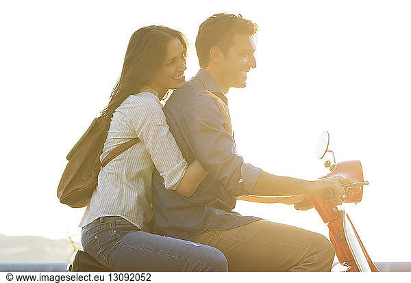 Side view of couple riding on motor scooter against clear sky on sunny day