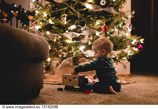 Side view of cute baby boy playing with toy truck by Christmas tree at home