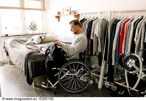 Side view of disabled mature man packing tennis racket in bag while sitting on wheelchair at home