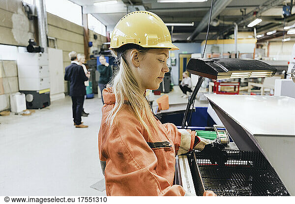 Side view of female blue-collar worker in hardhat using machinery in factory