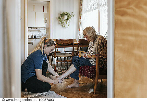 Side view of female care assistant helping senior woman wearing socks while sitting at home