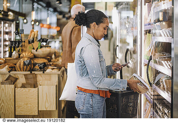 Side view of female customer photographing package while standing at grocery store