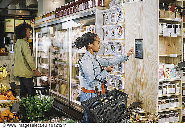 Side view of female customer using digital tablet mounted on wall at grocery store