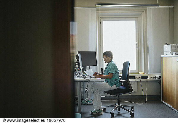 Side view of female doctor checking medical record while sitting at desk in office