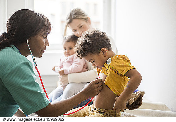 Side view of female doctor examining boy's heartbeat with stethoscope while mother sitting and daughter in clinic