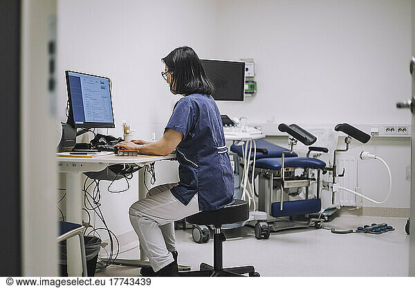 Side view of female doctor using desktop PC sitting at desk in medical clinic