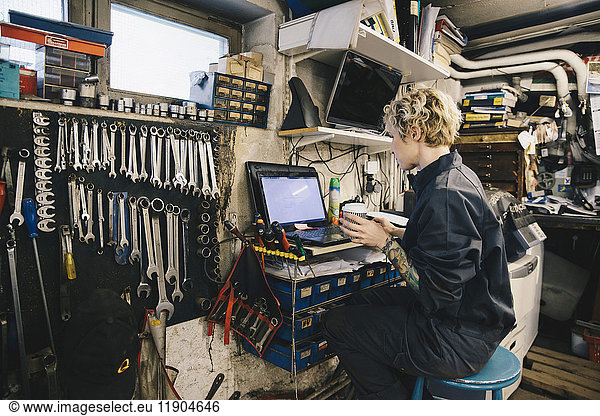 Side view of female mechanic holding disposable cup while using laptop at auto repair shop