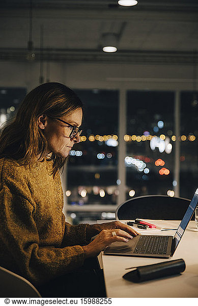 Side view of female professional working late while using laptop at illuminated desk in office