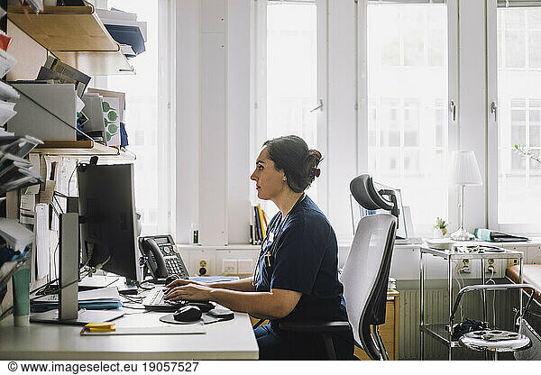Side view of focused female healthcare worker using computer while sitting on desk in clinic