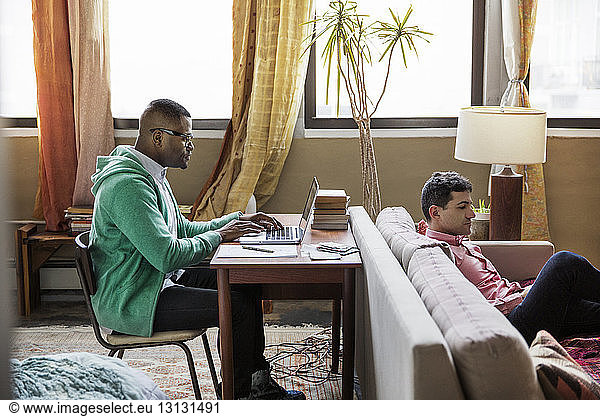 Side view of gay man using laptop at table while partner sitting on sofa