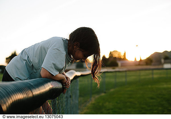 Side view of girl bending over fence at playground during sunset