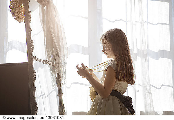 Side view of girl looking at necklace while standing by mirror at home