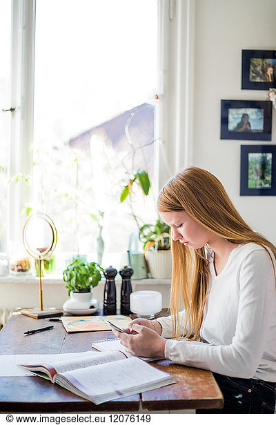 Side view of girl using smart phone while sitting with books at home