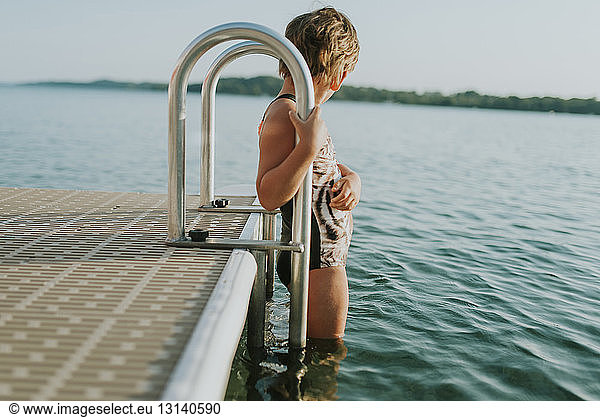 Side view of girl wearing swimwear while standing on ladder in lake against sky