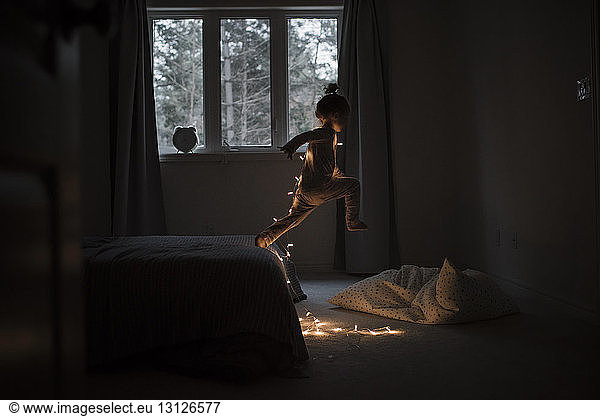 Side view of girl with illuminated string lights jumping from bed at home